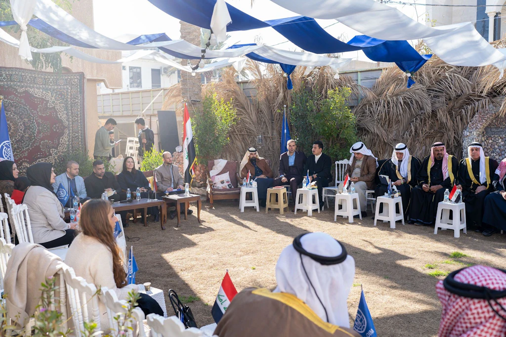 Representatives and tribal leaders seated in a circle under blue-and-white canopies, Iraqi and IOM flags in the foreground.IOM Iraq has confirmed the usage rights and the consent of the people shown in writing. Please use the caption and copyright line exactly as given.